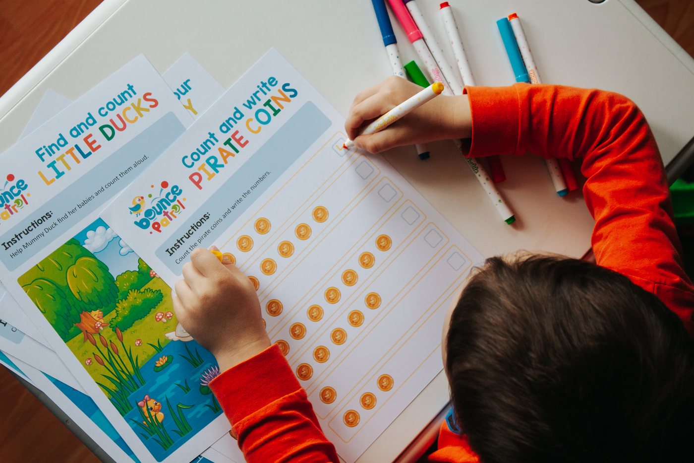 A child counting pirate coins on a Bounce Patrol worksheet, with more sheets and a pile of markers on the table